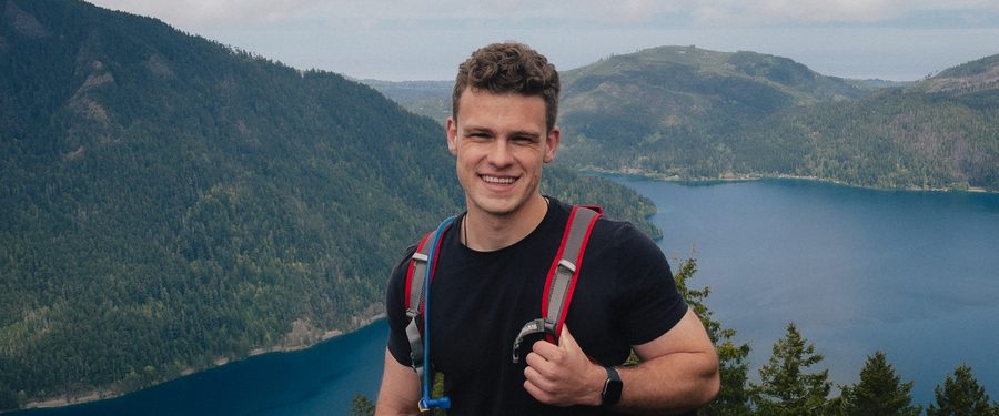 Jacob Haydel standing on a mountain overlook, backpack on, lake and forested hills behind him